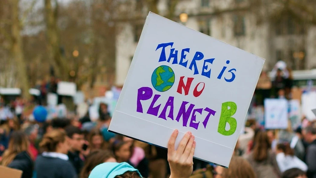 Tłum podczas protestu środowiskowego, gdzie ktoś unosi plakat z hasłem 'There is no Planet B', podkreślającym znaczenie ochrony środowiska. W tle widać rozmazane sylwetki ludzi.