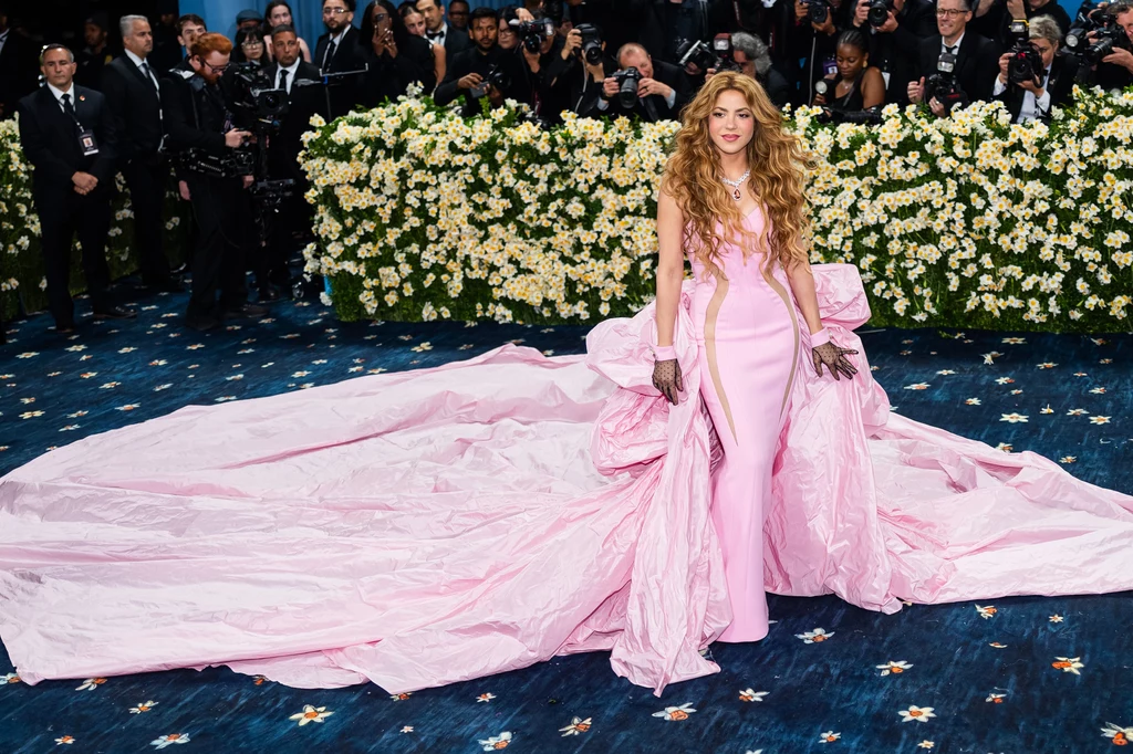 Shakira podczas Met Gali 2025 Osoba w eleganckiej, różowej sukni z długim trenem stoi na tle dekoracji kwiatowej. Dookoła znajduje się tłum fotografów, którzy uwieczniają ten moment. Stylizacja i otoczenie sugerują obecność na ekskluzywnym wydarzeniu.