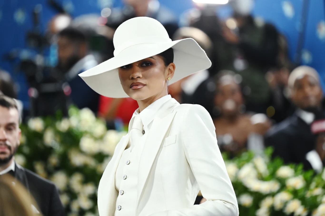 Kobieta ubrana w biały elegancki strój i duży kapelusz, stoi na tle tłumu ludzi. Jej wyraz twarzy jest spokojny, otoczenie wydaje się szykowne, wskazując na oficjalne wydarzenie.
