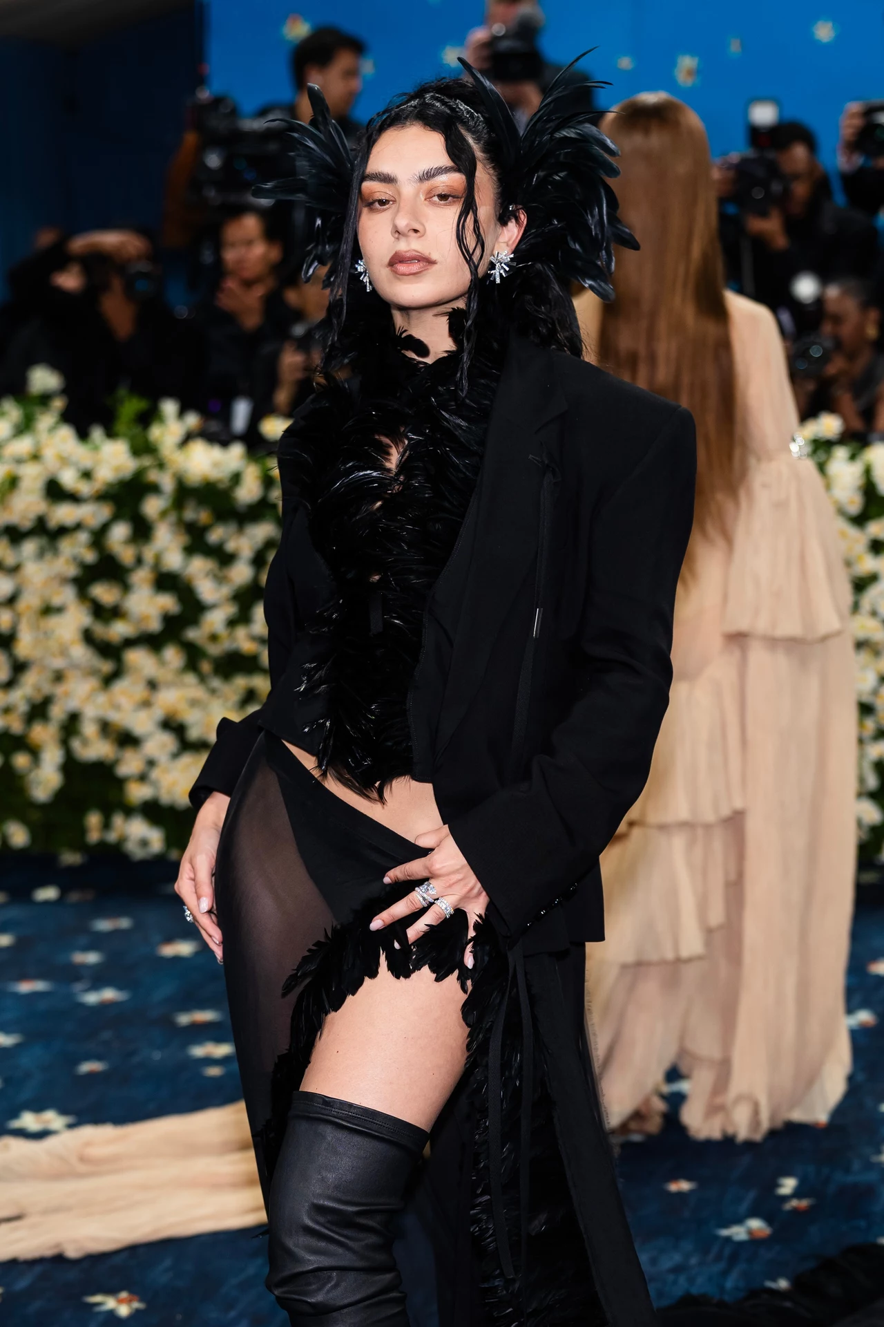Charli xcx na czerwonym dywanie Met Gali 2025 Kobieta w czarnej kreacji z piórami, stojąca na tle kwiatowej aranżacji na niebieskim dywanie. Na sobie ma elegancki płaszcz oraz buty o wysokich cholewkach. W tle widać innych uczestników oraz fotografów.