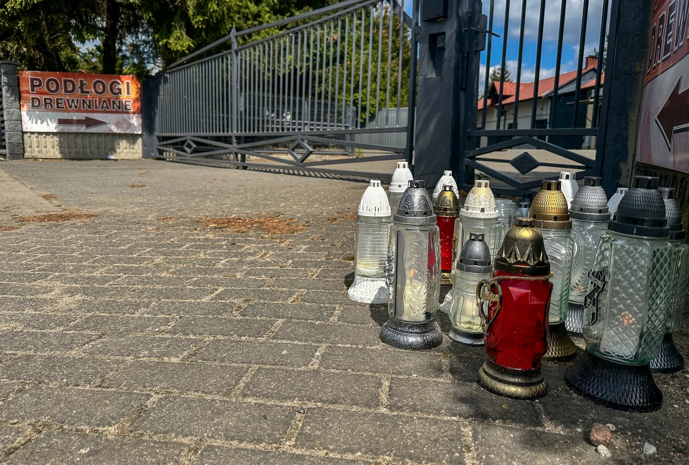 Znicze złożone ku pamięci nieżyjącej Mai z Mławy Znicze ustawione w kilka kręgów na chodniku przed zamkniętą bramą. W tle widoczny jest drewniany płot oraz tablica z napisem "Podłogi Drewniane".