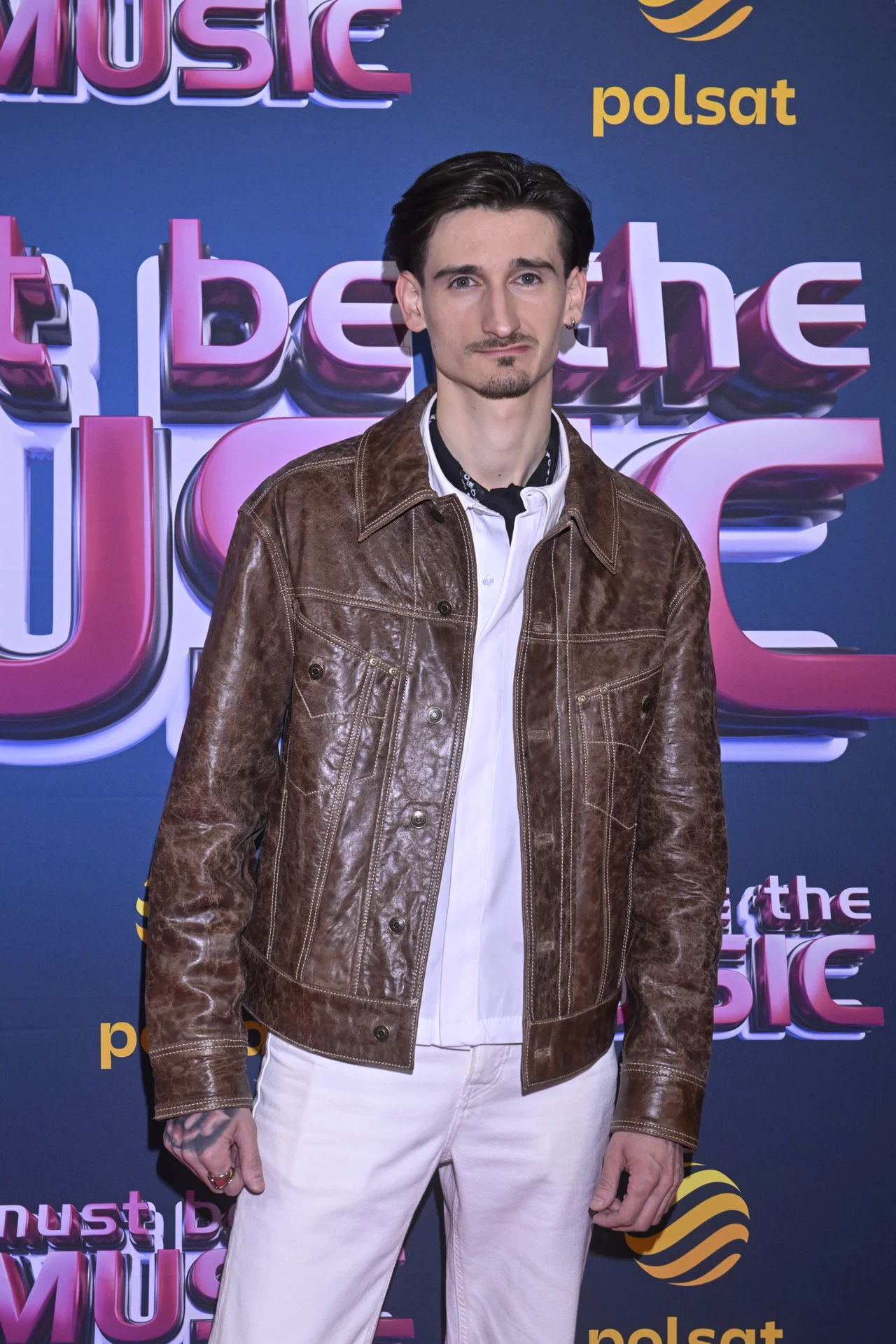 Dawid Dubajka wystąpi w finale "Must Be The Music" Mężczyzna ubrany w białą koszulę i białe spodnie, noszący brązową skórzaną kurtkę, stoi przed ścianką z napisem 'Must Be The Music' i logotypem Polsatu. Wygląda na podekscytowanego lub szczęśliwego, lekki uśmiech na twarzy.