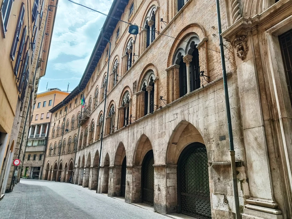 Różnorodna architektura miasta zachęca do skręcania w mniej uczęszczane uliczki Historyczny budynek z arkadami, zbudowany z cegły i kamienia, umiejscowiony w wąskiej uliczce, z włoską flagą powiewającą na jednej z fasad. Jasne niebo w tle, puste ulice tworzą spokojną atmosferę.