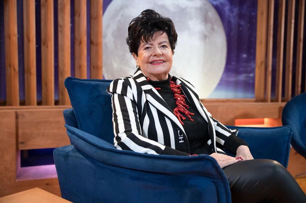 Osoba siedząca na niebieskim fotelu w studiu telewizyjnym, w tle duży obraz Księżyca.