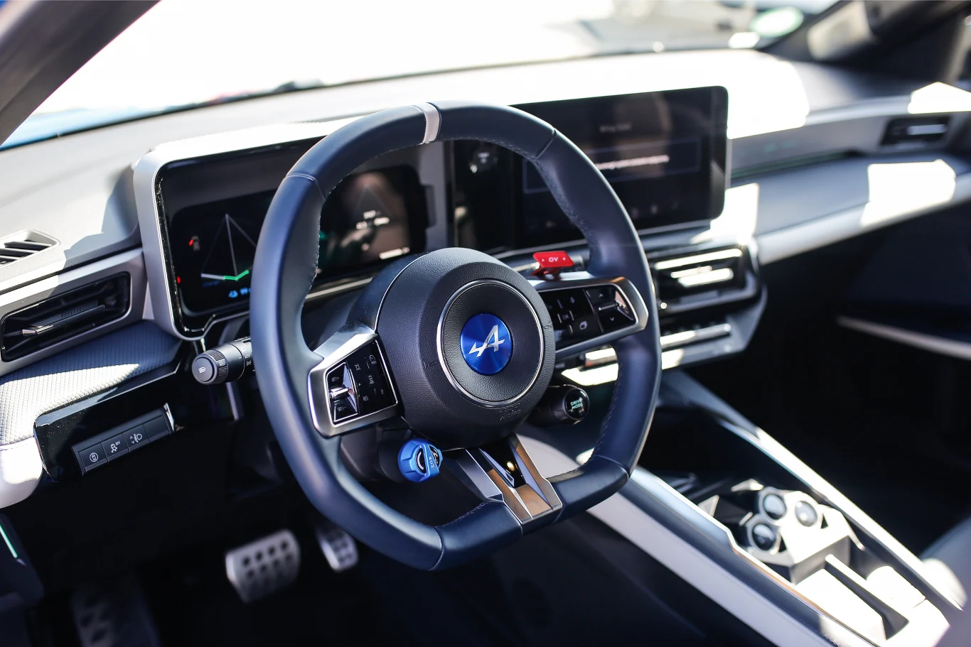 Wnętrze nowoczesnego samochodu, centralnie umieszczona kierownica z logo, wyświetlacze na desce rozdzielczej prezentujące cyfrowe wskaźniki oraz elementy sterujące na konsoli środkowej.