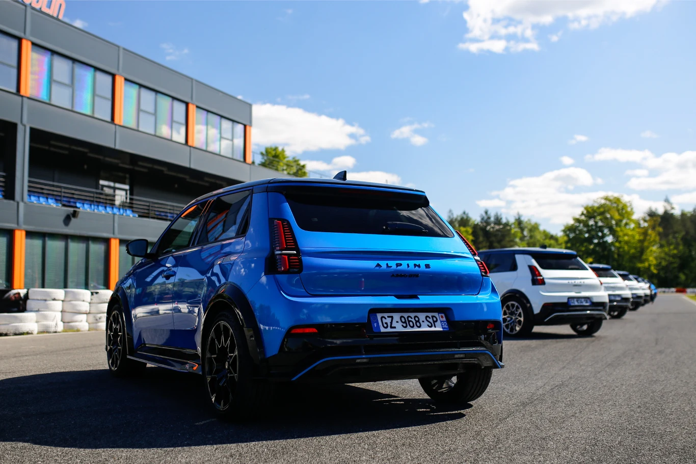 Niebieski samochód marki Alpine stoi na torze wyścigowym obok białego auta. W tle widoczny jest nowoczesny budynek z pomarańczowo-niebieskimi akcentami. Wyraźne światło dzienne i kilka białych opon po prawej stronie uzupełniają scenerię.