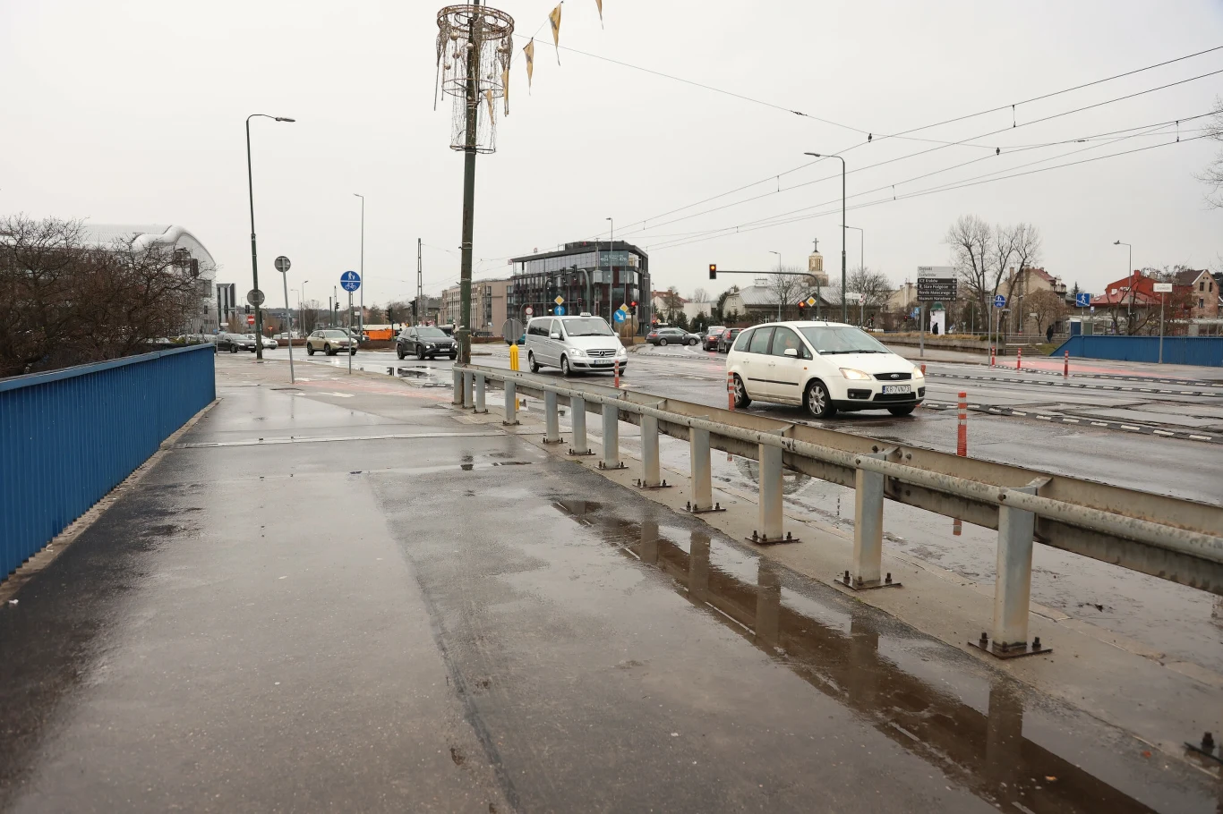 Ulica przy deszczowej pogodzie z kilkoma samochodami jadącymi po mokrym asfalcie, otoczona niskimi barierkami ochronnymi. W oddali widać budynki i znaki drogowe, co wskazuje na miejskie środowisko.