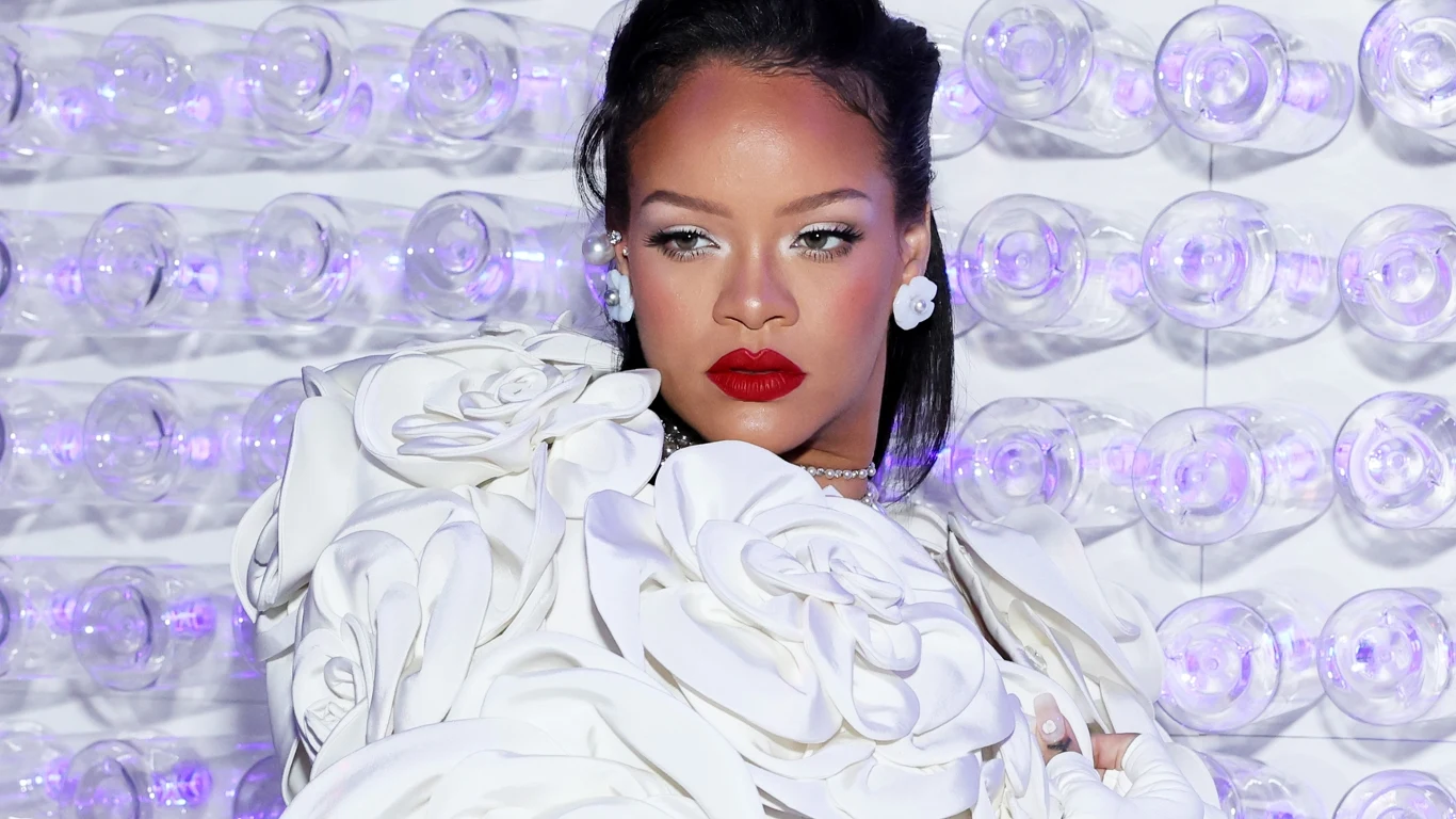 Ile posiadłości ma Rihanna? Kobieta ubrana w elegancką, białą suknię z kwiecistymi ozdobami, stojąca na tle ściany z przezroczystymi elementami oświetlonymi na fioletowo. Ma wyrazisty makijaż z czerwonymi ustami.