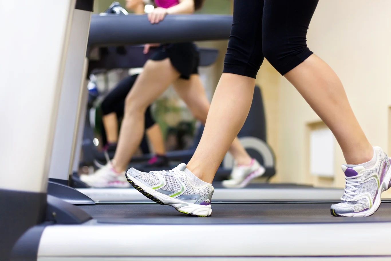 Osoby w legginsach i sportowych butach ćwiczące na bieżniach w siłowni, koncentrując się na nogach podczas biegania i chodzenia.