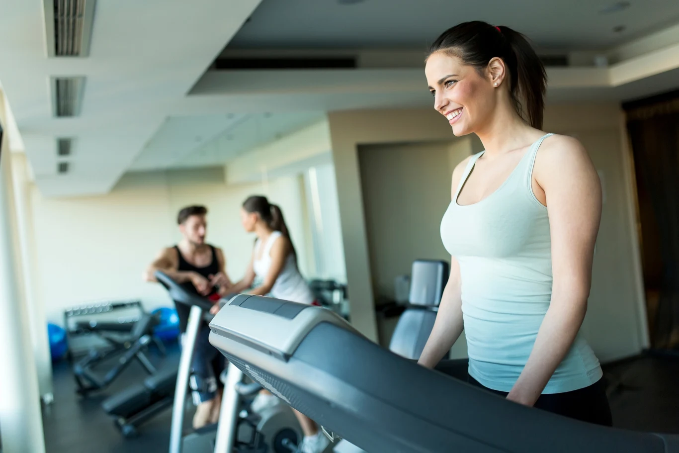 Kobieta ćwicząca na bieżni w nowoczesnej siłowni z dużymi oknami, w tle para rozmawiająca przy innych urządzeniach fitness.