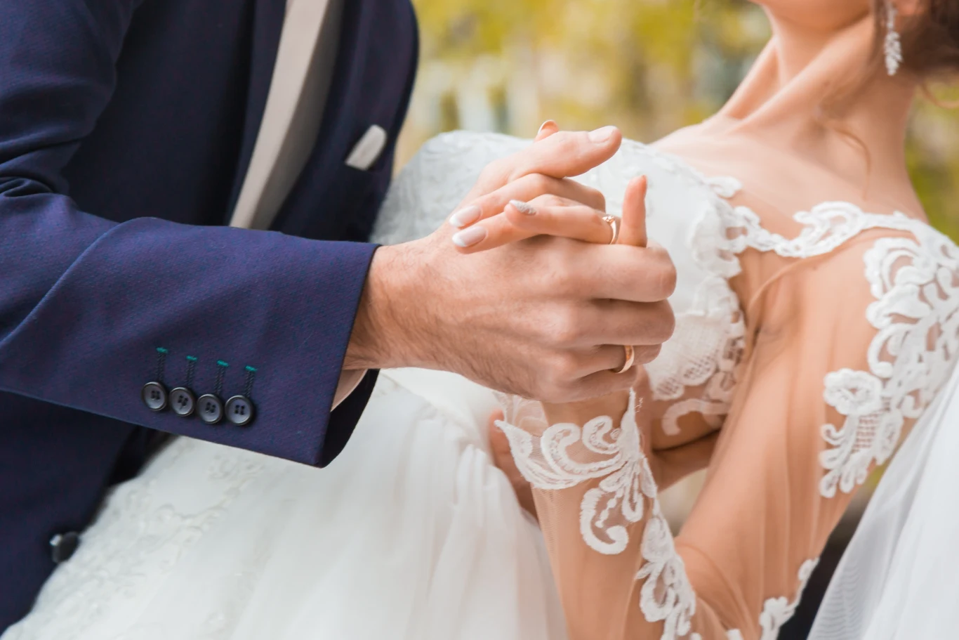 Para młoda w eleganckim tańcu, mężczyzna w granatowym garniturze trzyma rękę kobiety w białej koronkowej sukni ślubnej, zbliżenie na splecione dłonie, subtelne detale sukni i guzików marynarki.