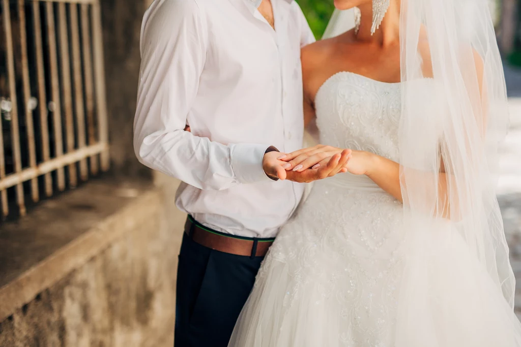 Zdejmowanie marynarki już na początku uroczystości weselnej jest złym pomysłem Para młoda w eleganckich strojach trzymająca się za ręce w dniu ślubu, z widoczną suknią ślubną i białą koszulą na tle kamiennego muru.