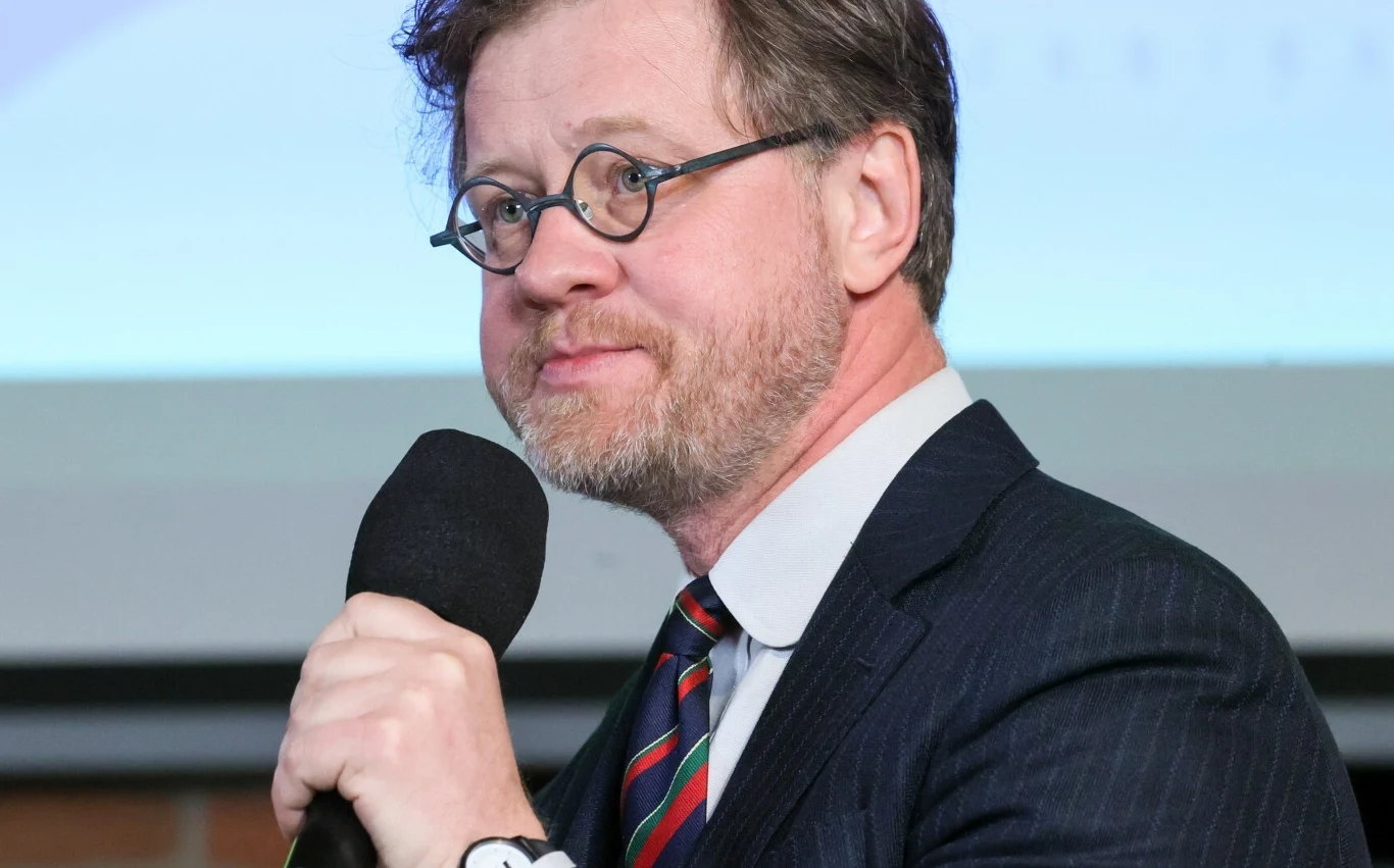 Znamy laureatów Konkursu Chopinowskiego. Wśród nich aż 13 Polaków Mężczyzna z okularami i brodą trzyma mikrofon, ubrany w elegancki garnitur i krawat, stoi na tle rozmytego ekranu.