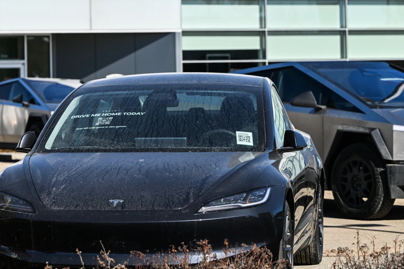 Czarny samochód elektryczny Tesla stoi na parkingu przed nowoczesnym budynkiem. Na szybie znajduje się napis. W tle widoczne są inne pojazdy, w tym futurystyczny samochód o nietypowym kształcie. Teren jest porośnięty suchą trawą.