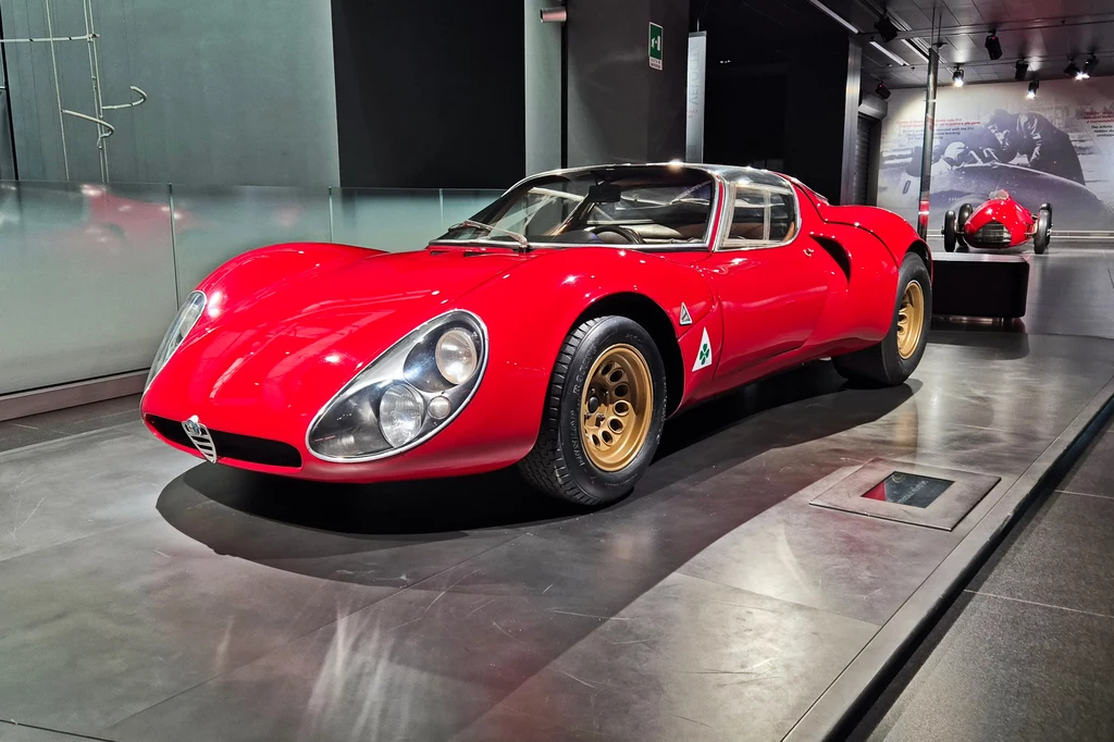 Alfa Romeo 33 Stradale zaprezentowano w 1967 roku Czerwony, klasyczny samochód sportowy zaprezentowany w nowoczesnym muzeum lub salonie, z dobrze oświetlonym wnętrzem. Auto ma aerodynamiczny kształt, duże przednie reflektory i złote felgi. W tle widać podobne auto oraz szklane ściany wystawowe.