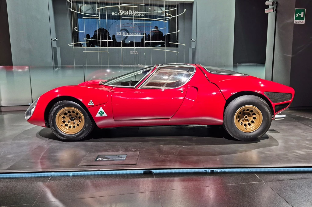 33 Stradale bazowało na wyścigowym Tipo 33 Elegancki czerwony samochód sportowy z klasycznym designem, eksponowany na wystawie w galerii. Posiada złote felgi i charakterystyczne logo na drzwiach. W tle nowoczesne wnętrze galerii i subtelne oświetlenie.