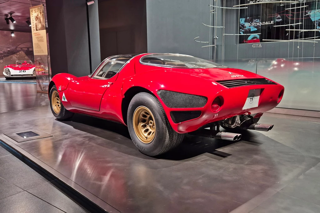 Powstało zaledwie 18 egzemplarzy Alfa Romeo 33 Stradale Czerwony sportowy samochód stojący na wystawie w muzeum motoryzacji. Pojazd wyróżnia się opływową sylwetką i sportowym designem. Tylna część samochodu posiada unikalne detale i liczne wloty powietrza. W tle inne eksponaty motoryzacyjne.