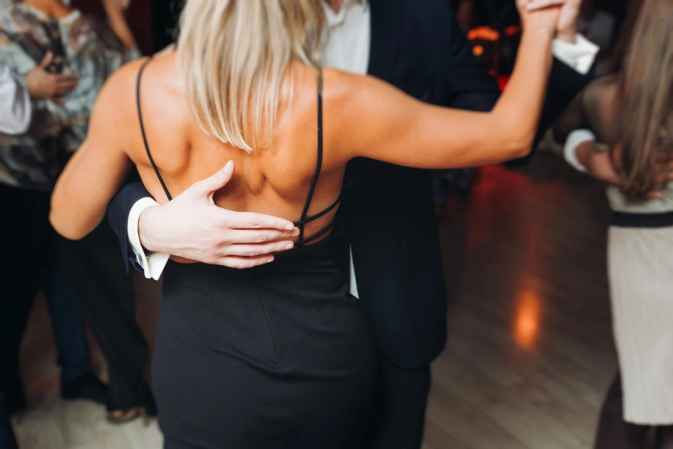 Taniec towarzyski w eleganckim otoczeniu, para tańczy blisko siebie, kobieta w czarnej sukni i mężczyzna w garniturze trzymają się za ręce, tworząc romantyczną atmosferę.