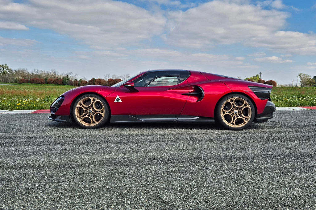 Alfa Romeo 33 Stradale Sportowy samochód w kolorze czerwonym na torze wyścigowym, z dynamiczną linią i złotymi felgami, na tle błękitnego nieba i zieleni krajobrazu.