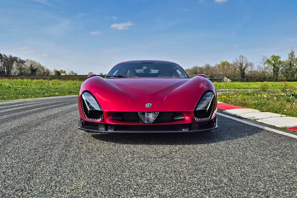 Alfa Romeo 33 Stradale Elegancki czerwony samochód sportowy o nowoczesnym designie stoi na asfalcie toru wyścigowego, otoczony zielenią i niebem z delikatnymi chmurami.