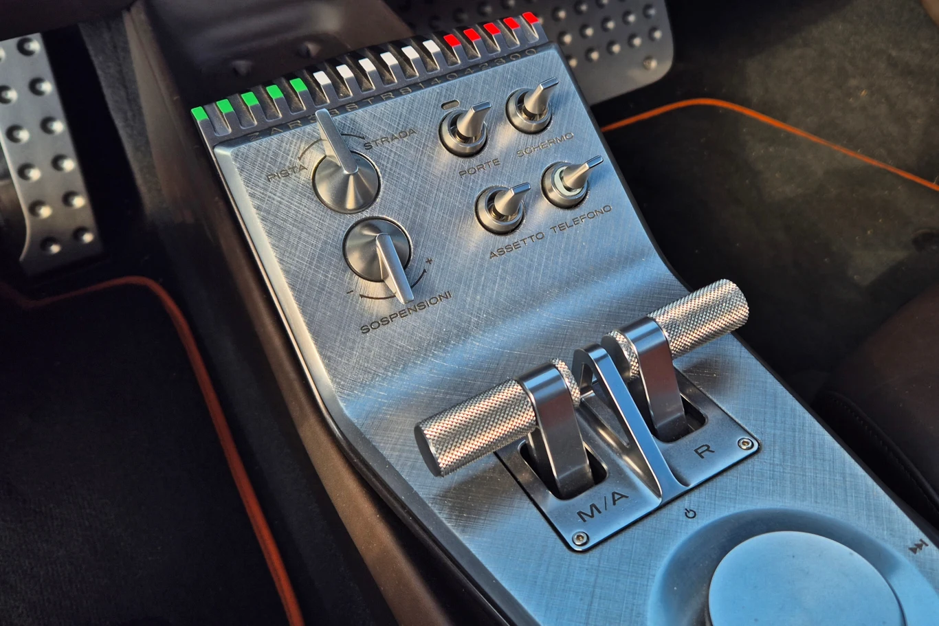 Panel sterowania w eleganckim samochodzie z metalowymi przełącznikami i dźwigniami, otoczony aerodynamiczną deską rozdzielczą. W tle widoczne pedały pokryte metalowymi nakładkami.