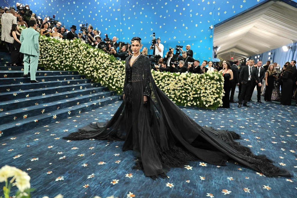 Dua Lipa na Met Gali 2025 Osoba w eleganckiej czarnej sukni z długim trenem stoi na schodach pokrytych niebieskim dywanem wśród licznie zebranych gości. W tle widać ścianę ozdobioną kwiatami oraz licznych fotografów i uczestników wydarzenia.