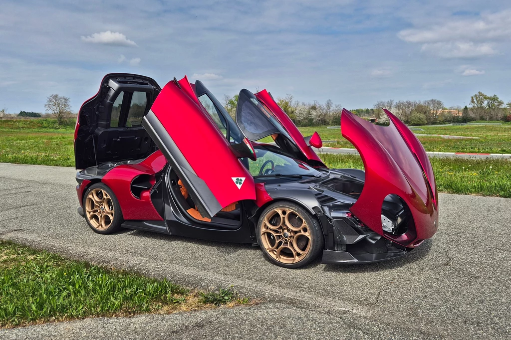 Alfa Romeo 33 Stradale po otworzeniu drzwi i klap wygląda obłędnie Wyjątkowy czerwony supercar z uniesionymi drzwiami i maską, stojący na asfaltowej drodze pośród zielonych pól. Samochód ma złote felgi i futurystyczny design podkreśla jego sportowy charakter.