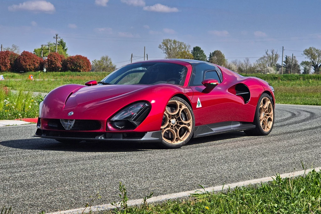 Alfa Romeo 33 Stradale Czerwony sportowy samochód jadący po krętej drodze, otoczony zielenią i jasno niebem z kilkoma chmurkami. Samochód ma nowoczesny, aerodynamiczny design i złote felgi, które podkreślają jego elegancję.