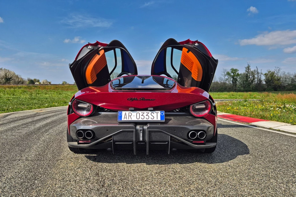 Alfa Romeo 33 Stradale Widok tylnej części sportowego samochodu Alfa Romeo z otwartymi drzwiami. Auto stoi na torze wyścigowym otoczonym zielenią, a w tle widać błękitne niebo z małą ilością chmur.