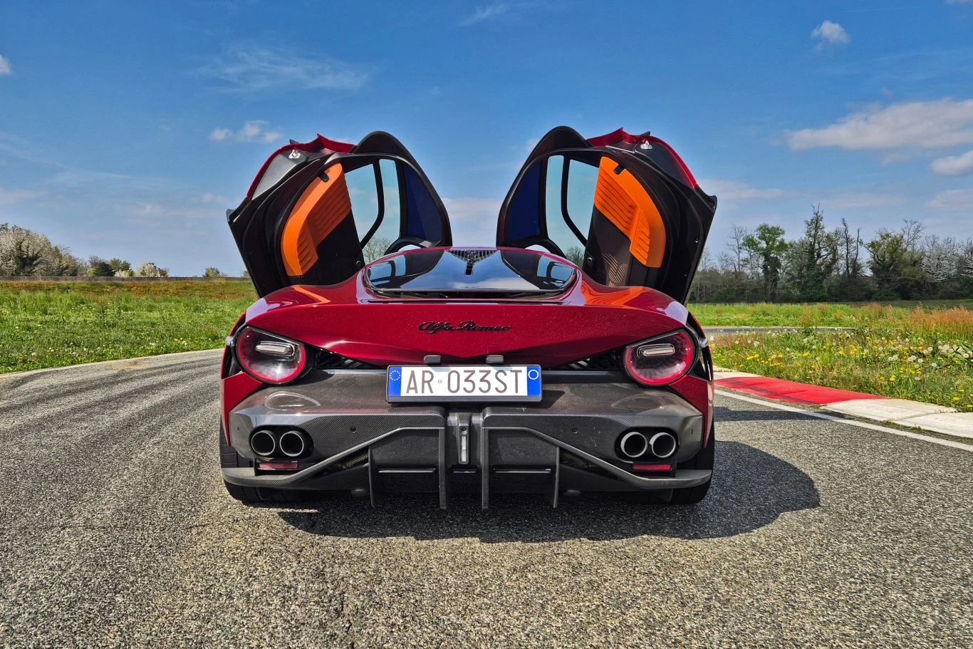 Widok tylnej części sportowego samochodu Alfa Romeo z otwartymi drzwiami. Auto stoi na torze wyścigowym otoczonym zielenią, a w tle widać błękitne niebo z małą ilością chmur.