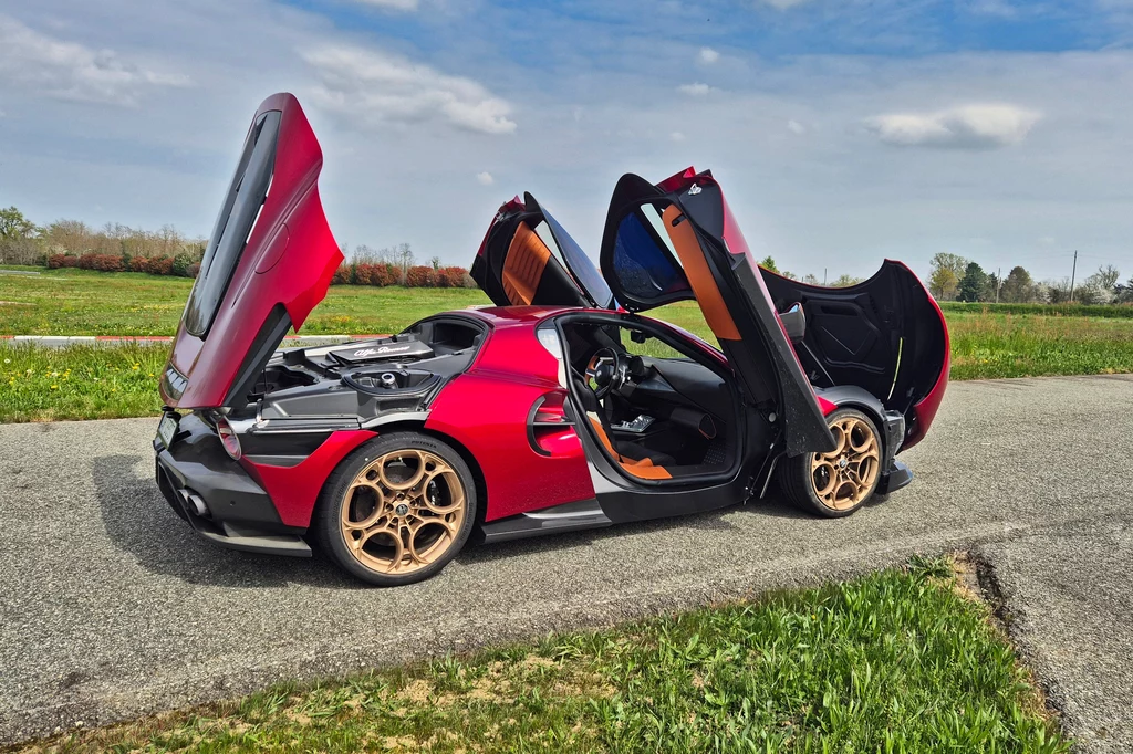 Alfa Romeo 33 Stradale Czerwony samochód sportowy z otwartymi drzwiami motylkowymi stoi na asfalcie przy polnej drodze. Na pierwszym planie widoczne są złote, stylizowane felgi i szczegóły mechaniczne silnika. W tle niebo z delikatnie zachmurzonymi chmurami.
