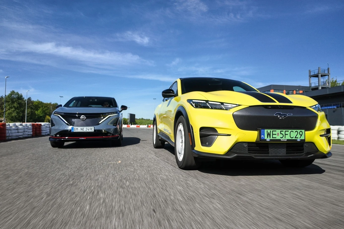 Dwa nowoczesne samochody sportowe na torze wyścigowym, żółty Ford Mustang Mach-E na pierwszym planie i ciemniejszy samochód w tle. W tle widać tor wyścigowy i jasne niebo z chmurami.