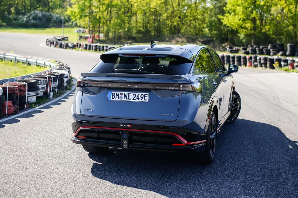 Nissan Ariya Nismo z zewnątrz wyróżnia się specjalnym pakietem aerodynamicznym Nismo (zderzaki, progi boczne, tylny spoiler) Samochód marki Nissan na torze wyścigowym, otoczony zielenią i barierami ochronnymi. Jasne, słoneczne niebo podkreśla nowoczesny design pojazdu, który jest zaparkowany w spokojnym otoczeniu.
