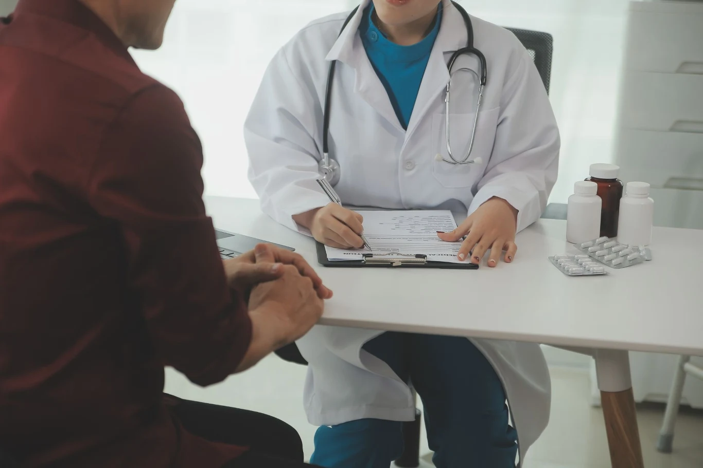 W niektórych przypadkach konieczna będzie wizyta u lekarza. Lekarz w białym fartuchu siedzi przy biurku z pacjentem, notując informacje na formularzu. Na stole widać leki w butelkach i blistry.