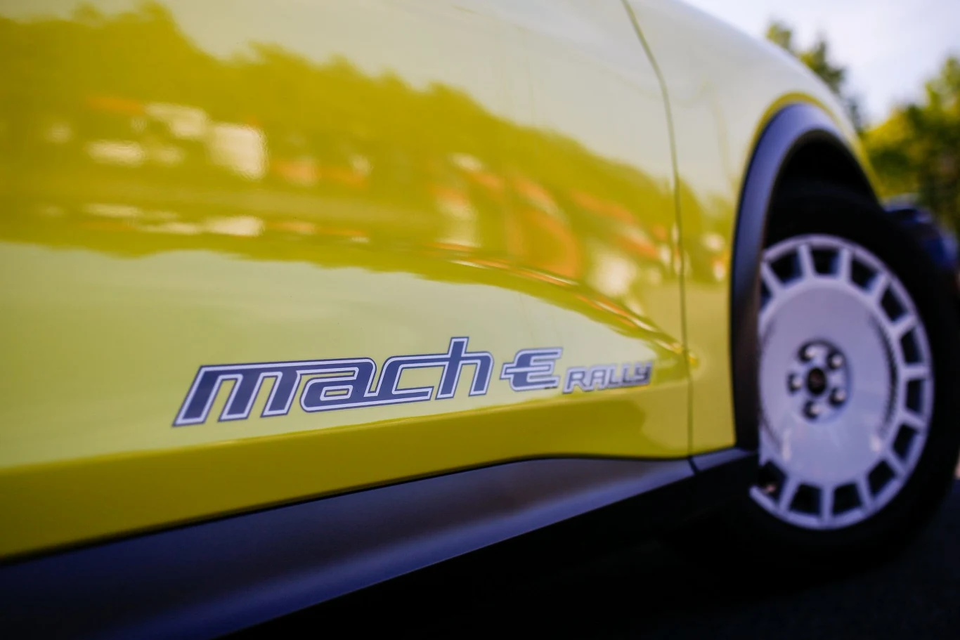 Bok żółtego samochodu z napisem "mach-e" obok charakterystycznego koła o sportowym wyglądzie. Auto jest w dynamicznym ujęciu, co podkreśla jego sportowy charakter.