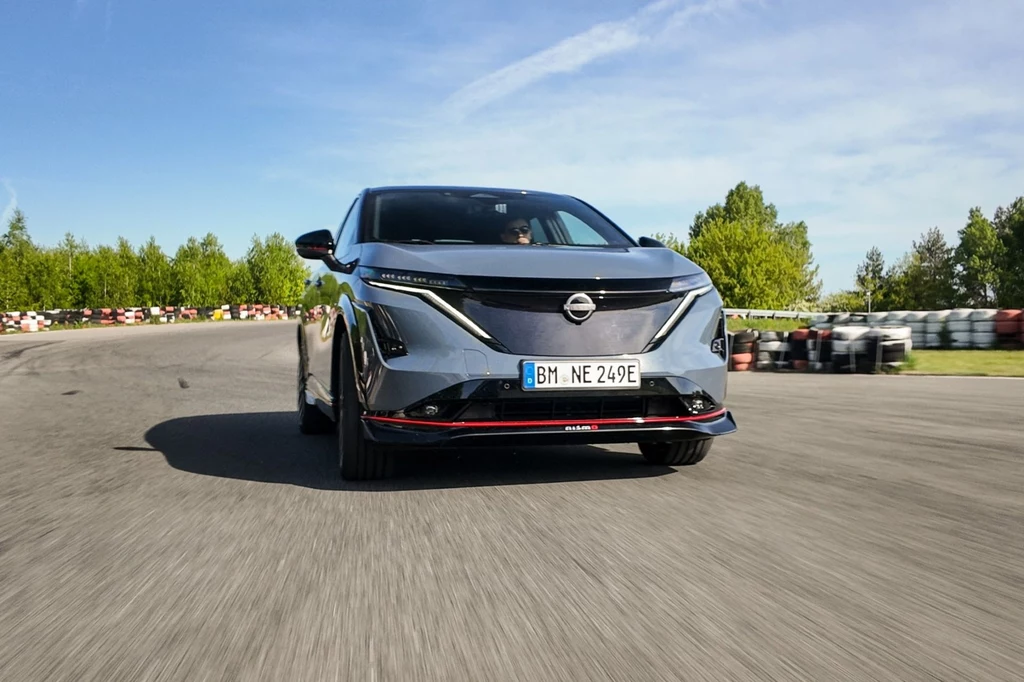 Nissan Ariya Nismo generuje 435 KM i 600 Nm. Nowoczesny samochód Nissan poruszający się po torze wyścigowym, otoczony zielenią i niebieskim niebem, z wyraźnie widocznym numerem rejestracyjnym.