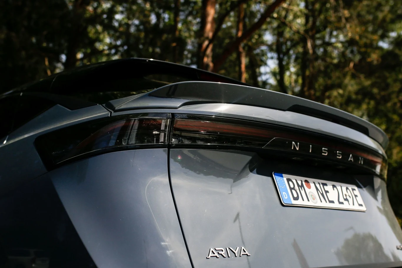 Tylne światła samochodu Nissan Ariya z widoczną tablicą rejestracyjną i emblematem marki. Samochód stoi na tle drzew, w świetle dziennym, podkreślając nowoczesny design pojazdu.