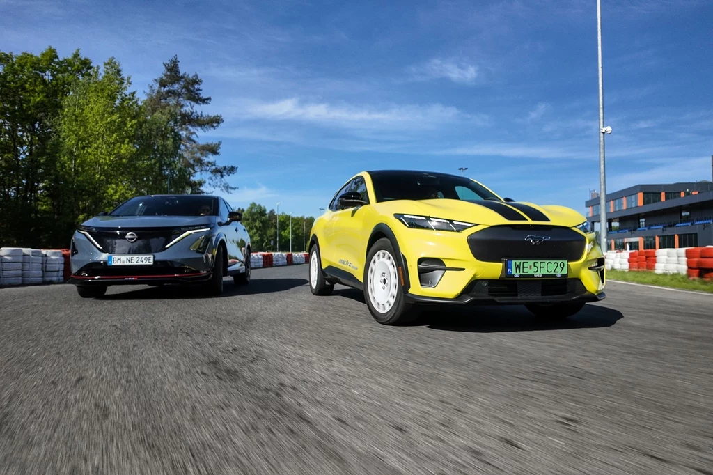 Nissan Ariya Nismo jest znacznie bardziej miękka niż Mustang Mach-E Rally. Dwa nowoczesne samochody elektryczne w dynamicznej jeździe na torze wyścigowym. Żółty SUV jadący z przodu po prawej, za nim czarny samochód o sportowym designie. W tle zieleń drzew oraz niebieskie niebo.