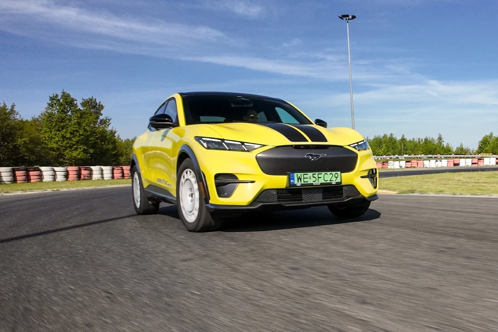 Mustang Mach-E Rally, jak na Forda przystało, prowadzi się świetnie. Delikatnym rozczarowaniem jest ciągle za lekko pracująca kierownica, nawet w topowym trybie jazdy - Untame. Żółty samochód sportowy o nowoczesnym designie na torze wyścigowym. W tle widoczne są barierki ochronne i drzewa, a pogodne niebo dodaje dynamiki scenie.