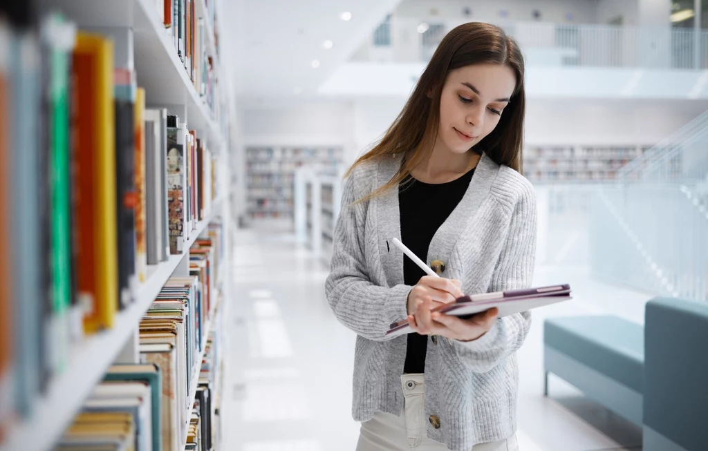 Panna to urodzony naukowiec Młoda kobieta w swetrze przegląda notatki w notesie stojąc pomiędzy regałami w nowoczesnej bibliotece. Światło we wnętrzu jest jasne i rozproszone, co tworzy spokojną atmosferę sprzyjającą nauce i refleksji.