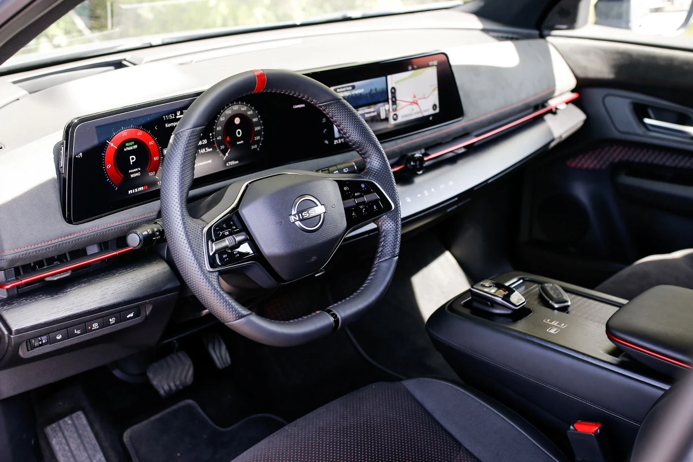 Nowoczesne wnętrze samochodu marki Nissan z dużym, szerokim ekranem na desce rozdzielczej. Centralne miejsce zajmuje wielofunkcyjna kierownica z logo. Obok widoczna jest konsola z kontrolerem biegów i różnymi przyciskami.