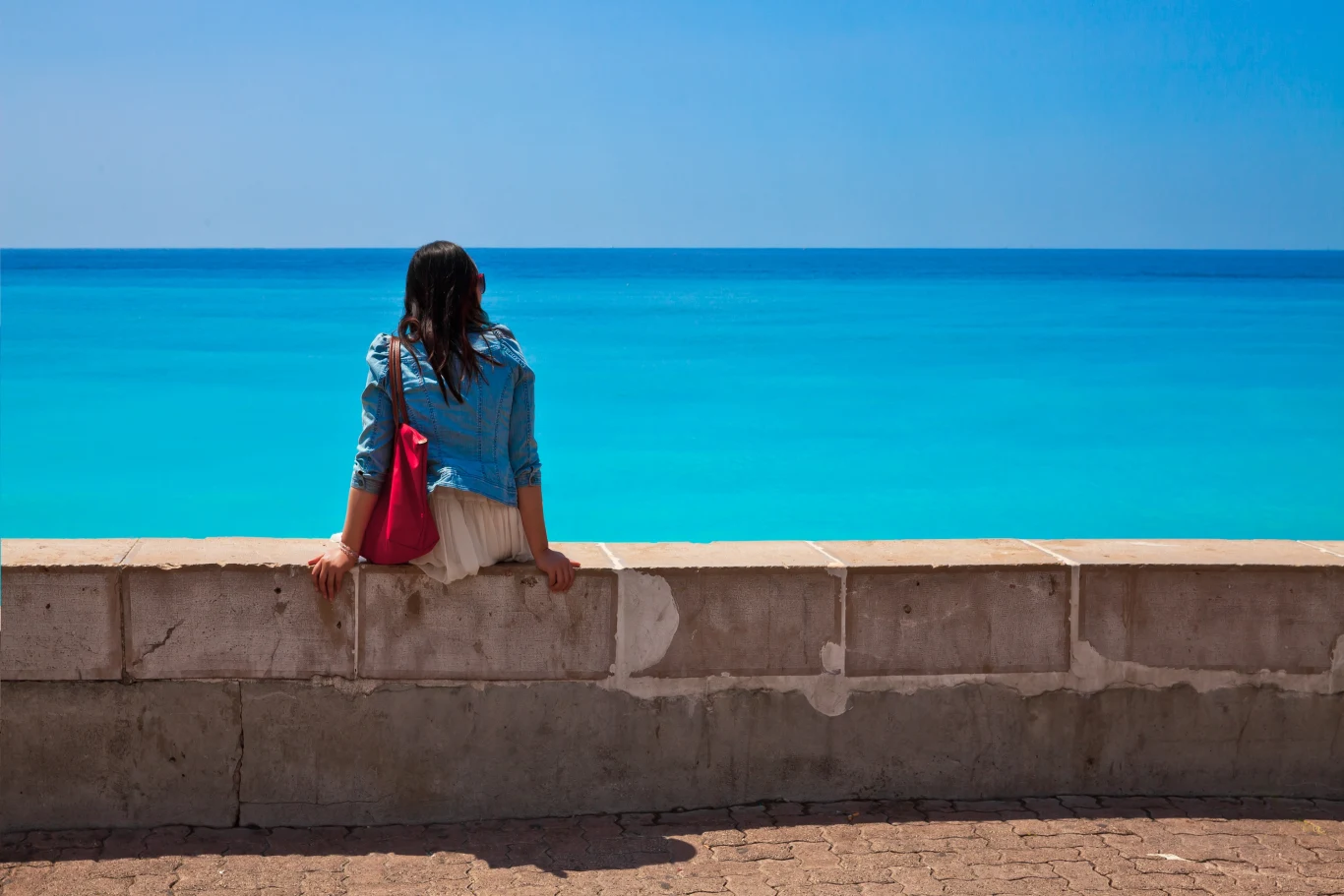 Osoba siedząca na murku z widokiem na turkusowe morze. Na sobie ma niebieską kurtkę i różową torbę na ramieniu, a niebo jest jasnoniebieskie i bezchmurne.