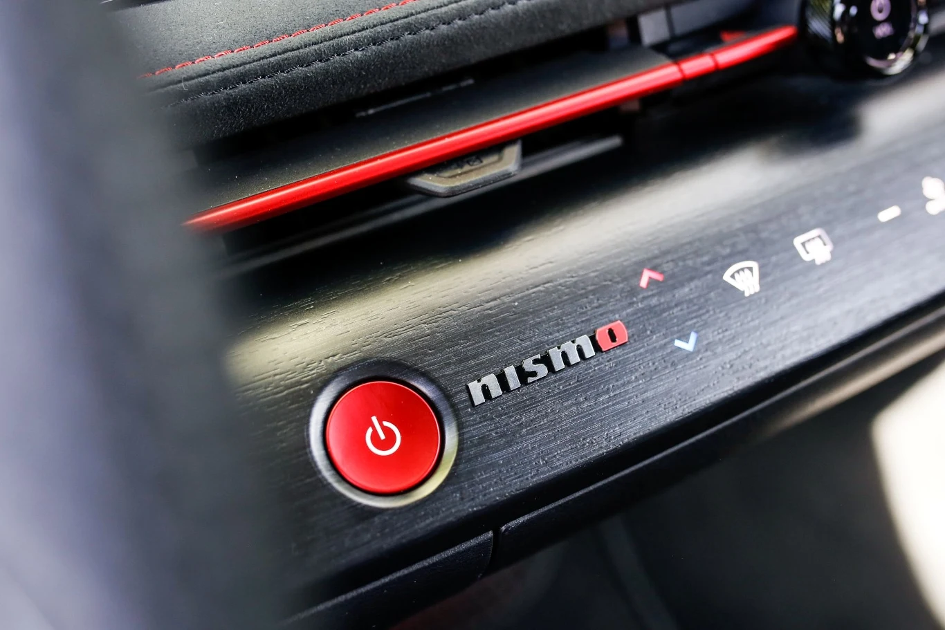 Przycisk startu w samochodzie z logo Nismo na desce rozdzielczej, wykończony eleganckim materiałem z czerwonymi akcentami.