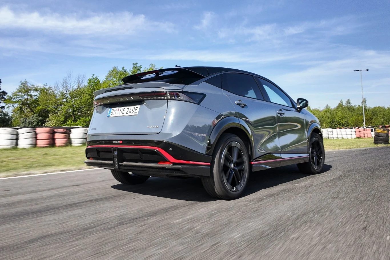 Widok na nowoczesny samochód elektryczny w ruchu na torze wyścigowym otoczonym przez opony. Pojazd w srebrnym kolorze z czarnymi detalami i aerodynamicznym designem.