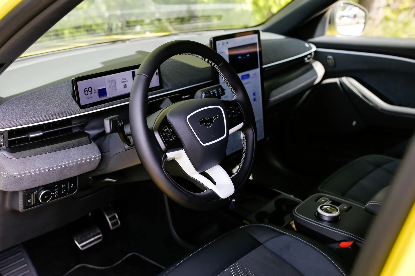 Wnętrze nowoczesnego samochodu z logiem Mustanga na kierownicy. Centralny wyświetlacz dotykowy i cyfrowy panel kontrolny. Charakterystyczne elementy designu z czarnymi wykończeniami w żółtym pojeździe.