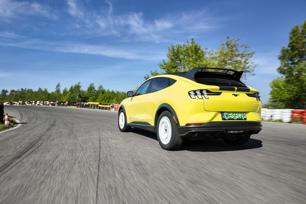Na rozpędzenie się do 100 km/h Ford Mustang Mach-E Rally potrzebuje 4,1 sekundy. Podobnie jak w Ariyi Nismo prędkość maksymalna wynosi 200 km/h. Żółty samochód elektryczny sportowy na torze wyścigowym podczas dynamicznej jazdy. W tle otoczony zielenią, niebieskim niebem i czerwono-białymi barierkami ochronnymi.