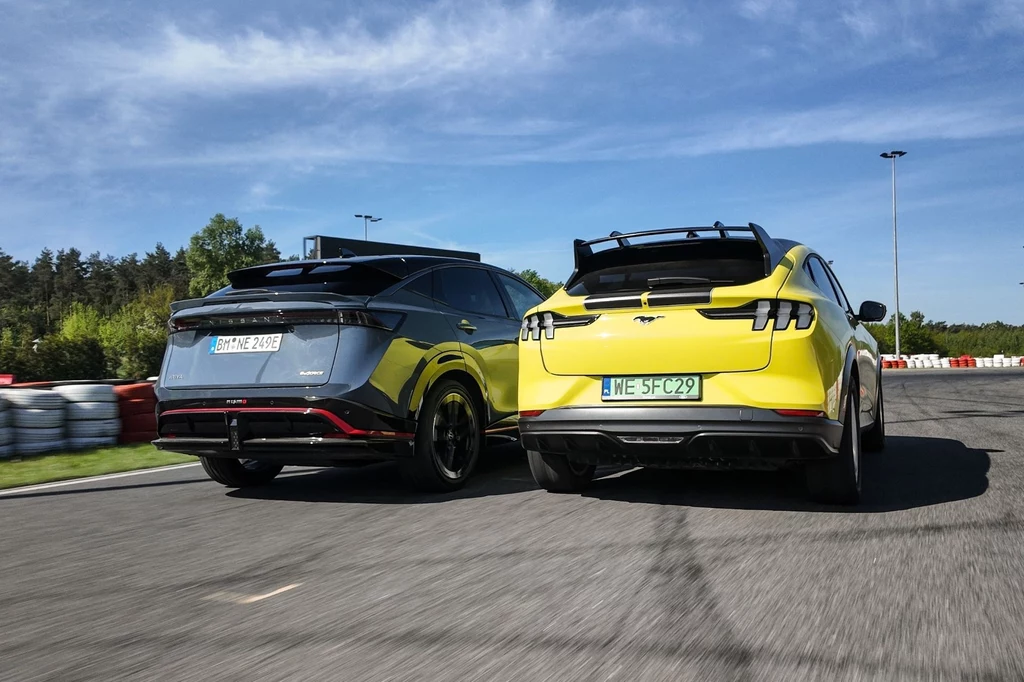Ford Mustang Mach-E Rally jest mocniejszy, ma lepszy czas do setki, a także większy akumulator i deklarowany zasięg. Dwa nowoczesne samochody sportowe na torze wyścigowym w ruchu. Żółty i czarny pojazd z aerodynamicznymi spojlerami, poruszające się obok siebie na tle zielonego krajobrazu i niebieskiego nieba z białymi chmurami.