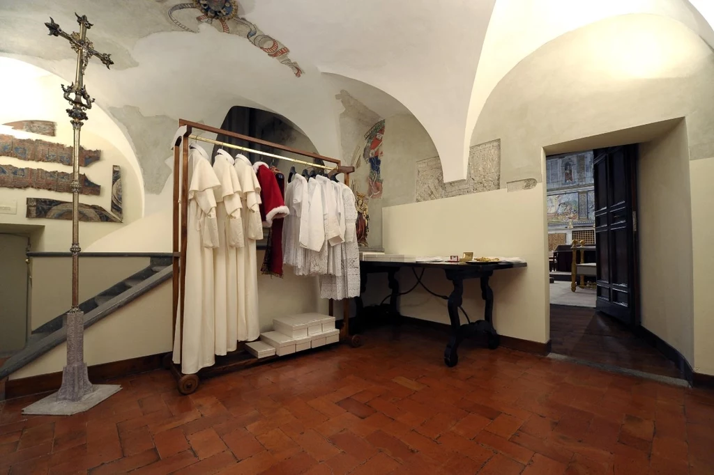 Na nowego papieża czekają trzy komplety strojów Pomieszczenie z zabytkowymi freskami na ścianach, w którym znajduje się wieszak z szatami liturgicznymi. Na ścianie widać barwne malowidła, a na podłodze terakotowe płytki. W tle otwarte drzwi prowadzące do kolejnego pomieszczenia.