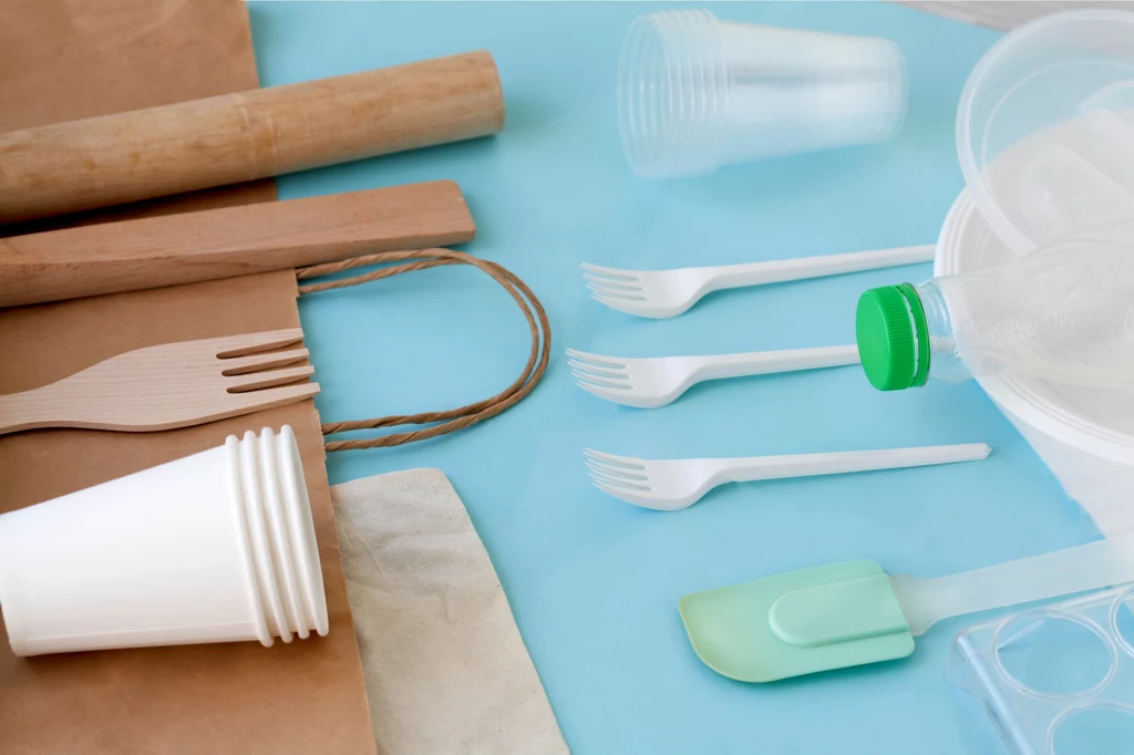 Świadomy wybór biodegradowalnych opcji zamiast plastikowych może pomóc w ratowaniu środowiska. Sztućce jednorazowe z plastiku i drewna, papierowe torby, plastikowe kubki i butelki oraz silikonowa szpatułka leżą na niebieskim tle, symbolizując różnorodność materiałów używanych w codziennych przedmiotach użytku jednorazowego.