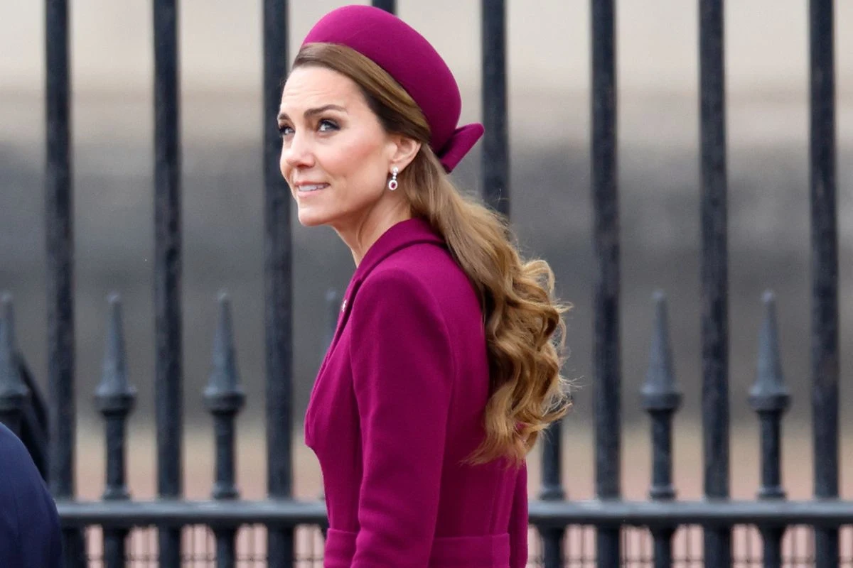 Księżna Kate zachwyciła wyborem koloru Kobieta w eleganckim bordowym stroju i nakryciu głowy, z długimi, falującymi włosami, stoi przed metalowym ogrodzeniem. Ma na sobie kolczyki.
